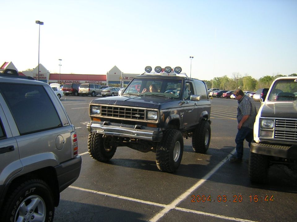 Lifted dark blue Ford Bronco II with roof lights parked in a lot next to a silver SUV and a white pickup