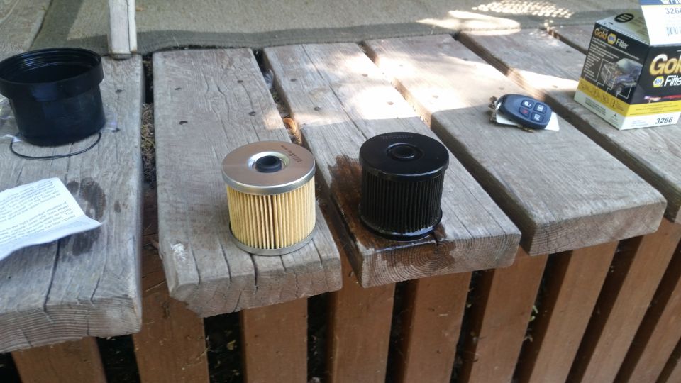 Two oil filters, a black cap, car keys, and a box of Gold Filter are on wooden planks