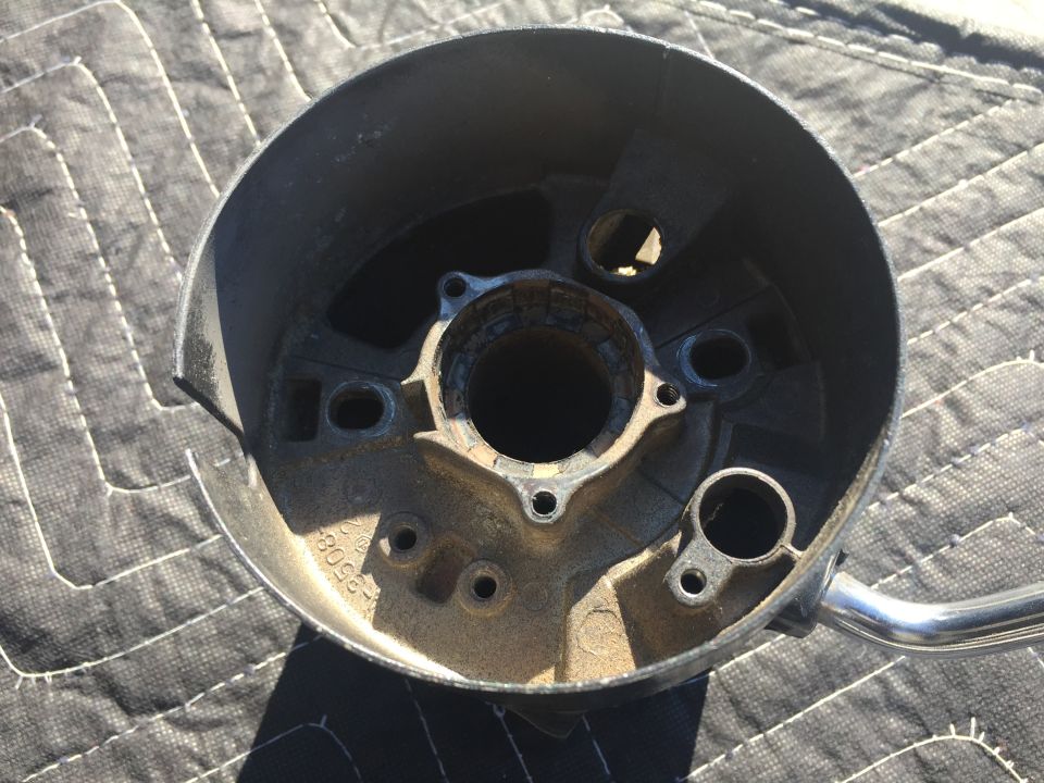 Close-up of a metal steering column housing with bolt holes and a central opening. Part number visible