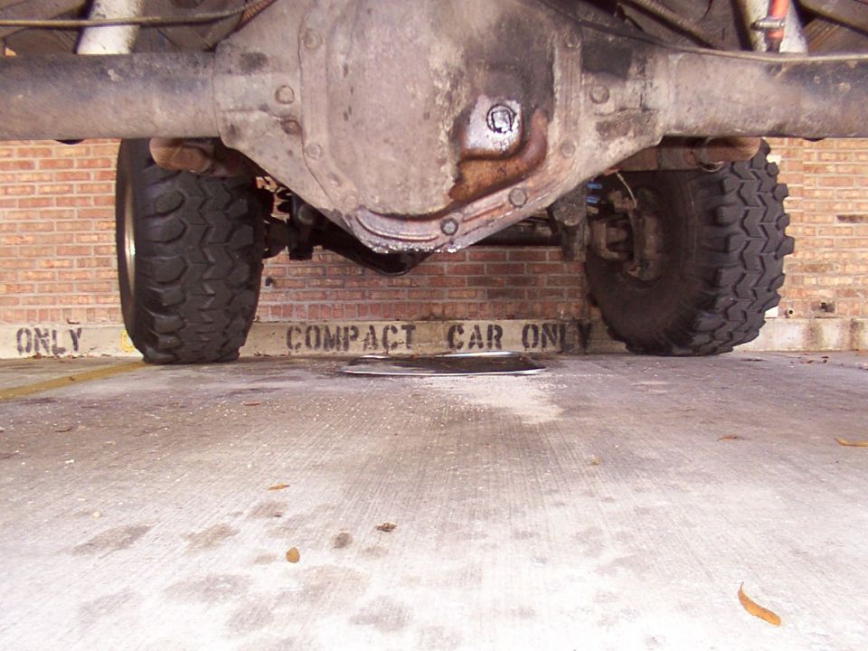 Rear axle and large tires of a truck, viewed from below. Brick wall with 'COMPACT CAR ONLY' text is visible