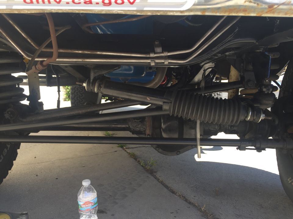 Underside view of a vehicle's steering and suspension components, with a bottle of water on the ground