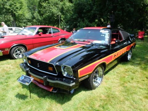 1971 Ford Ranchero - Black 1977 Ford Mustang Cobra II with red stripes and 'COBRA' decal on the side, parked on grass.