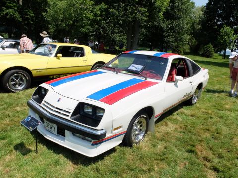 1971 Ford Ranchero - A white 1977 Pontiac Firebird Trans Am with red and blue racing stripes is displayed on a grassy field next to a yellow car.