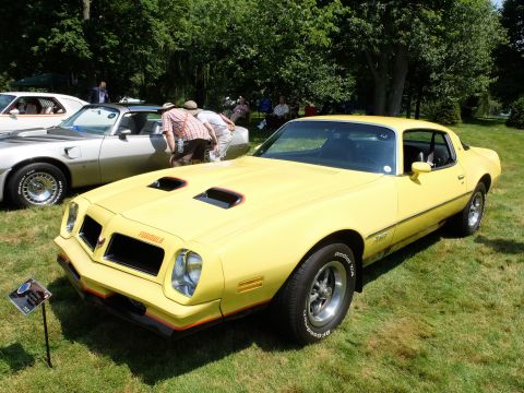 1971 Ford Ranchero - A yellow Pontiac Firebird Formula is displayed outdoors next to another car, with people gathered around.