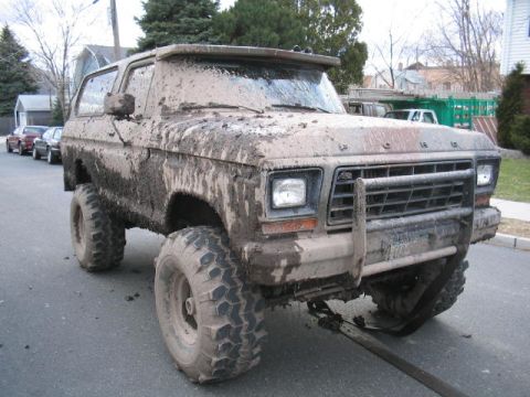 1979 Ford Bronco - This heavily mud-caked 1979 Ford Bronco is parked on a street, with other vehicles in the background.