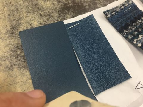 1978 Ford Bronco - Hand holds two textured dark-blue vinyl interior material samples against a worn workbench, blue tweed cloth swatch at top right.