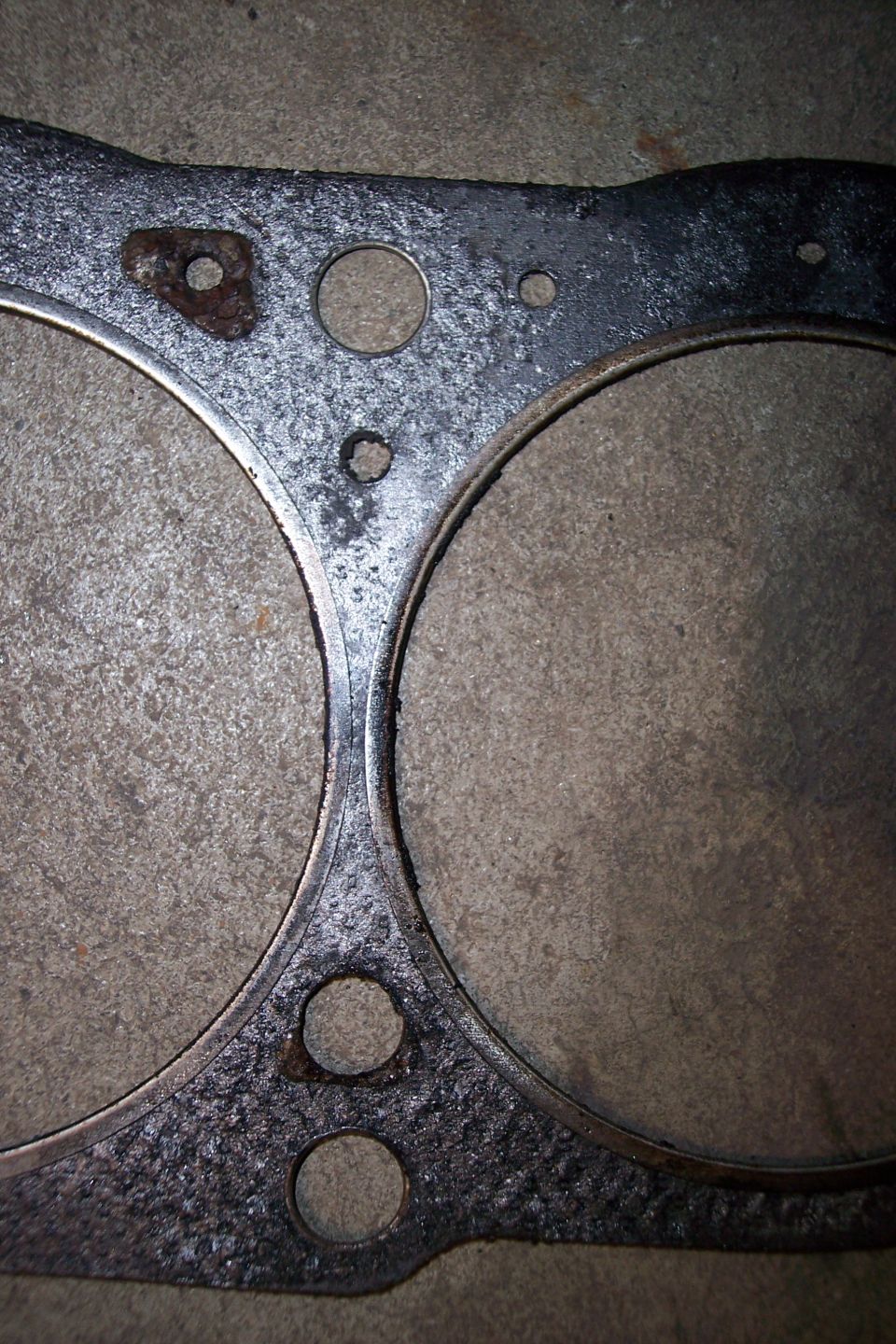 Engine gasket with two large circular openings and several smaller bolt holes, showing rust and wear