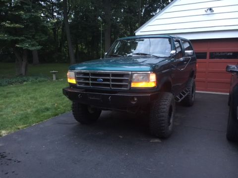1996 Ford Bronco - A green Ford Bronco is parked in front of a garage with its headlights on.