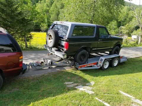 1996 Ford Bronco - A black Ford Bronco is loaded onto a U-Haul trailer, hitched to a red SUV, in a grassy, wooded area.