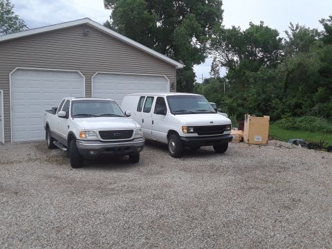 1999 Ford E250 - A white Ford E-Series van and a white Ford F-150 pickup truck are parked in a gravel driveway in front of a garage.