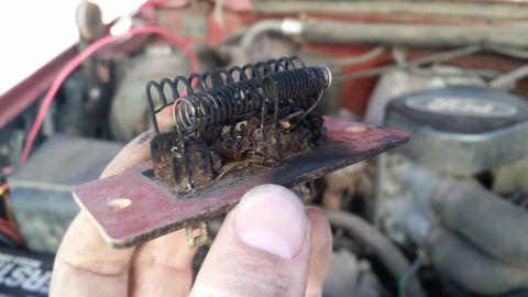 1985 Ford Bronco - A burnt blower motor resistor is held up for inspection, with the engine bay of a vehicle in the background.
