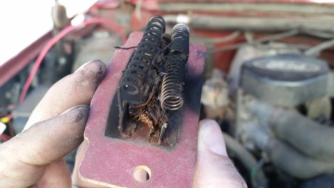 1985 Ford Bronco - A close-up of a damaged blower motor resistor, with the engine bay of a red vehicle visible behind it.