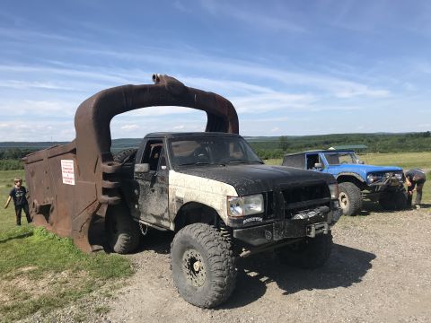 1989 Ford Bronco - Two off-road vehicles are parked outdoors on a sunny day, with one truck a massive, rusty metal attachment.