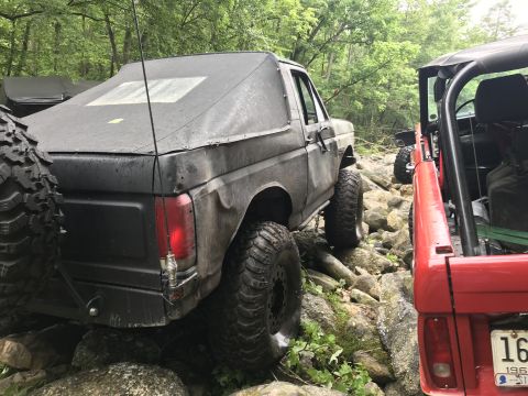 1989 Ford Bronco - A black 1989 Ford Bronco with a soft top is seen off-roading on a rocky path, with a red Jeep visible to its right.