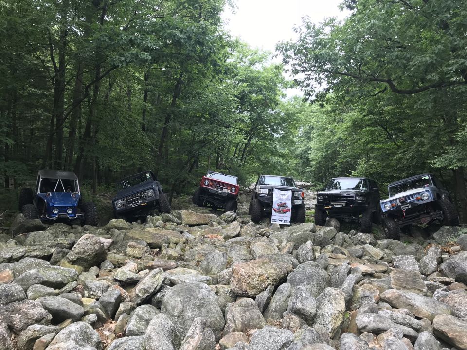 Five off-road vehicles navigate a rocky, wooded trail; one is blue, two are black, one is red, and one is dark blue