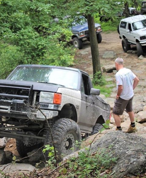 1989 Ford Bronco - Several off-road vehicles, including a Ford Bronco, tackle a challenging rocky terrain on a wooded trail.