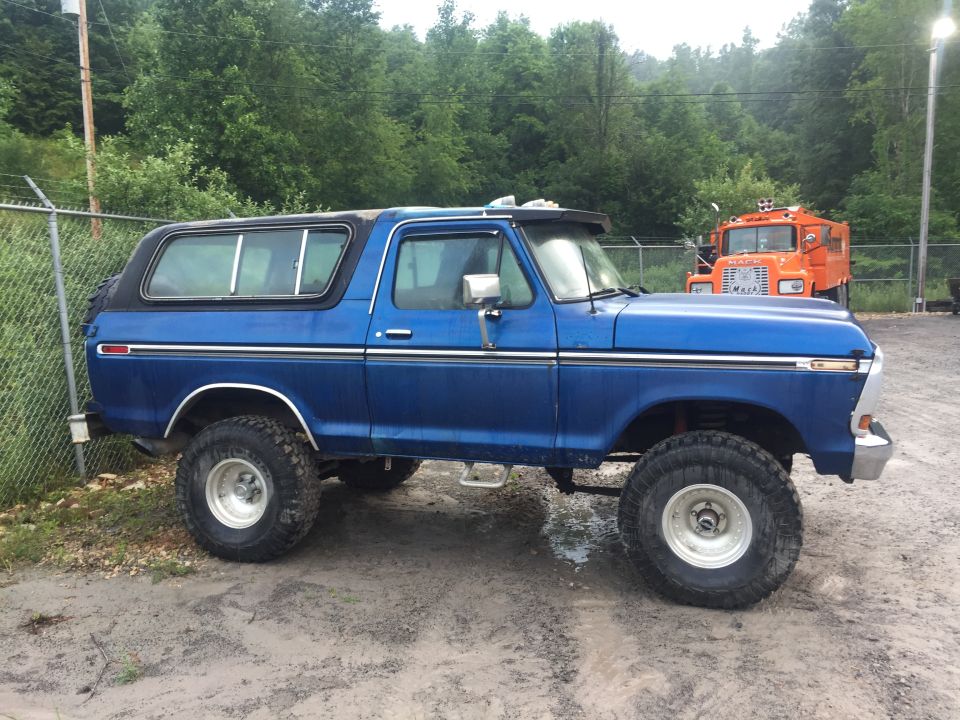 Blue 1978 Ford Bronco with a black hardtop, lifted suspension, and off-road tires. Parked on dirt next to a fence. An orange truck is in the background