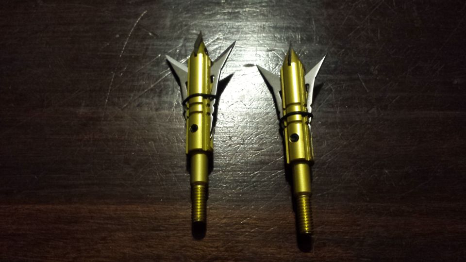 Two gold mechanical broadhead arrow tips with silver blades on a dark wooden table