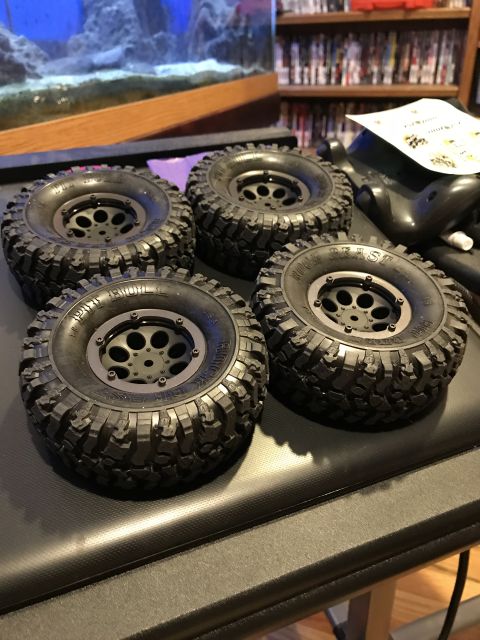 1989 Ford Bronco - Four black RC car tires with aggressive tread and multi-spoke wheels are displayed on a dark surface, with an aquarium in the background.