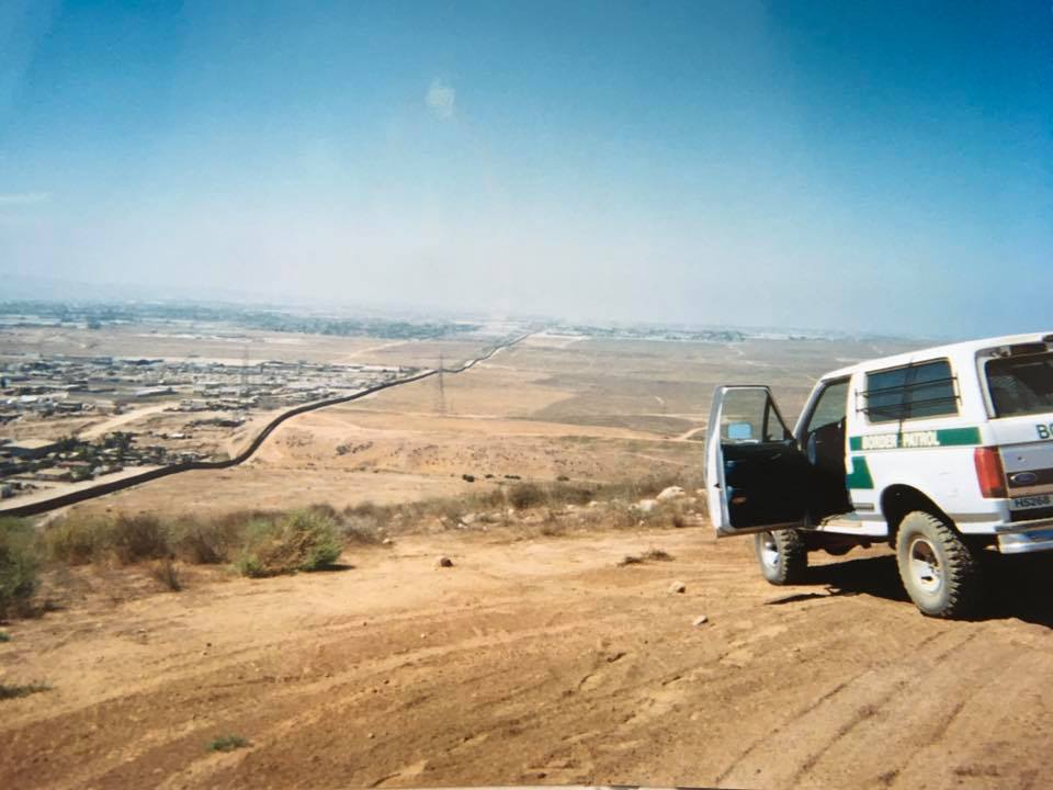 White 1996 Ford Bronco with open door parked on a hill overlooking a border wall and city.