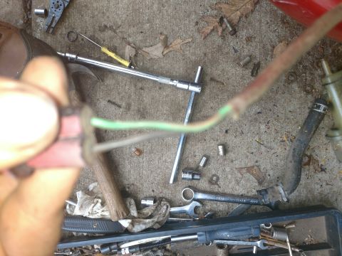 Hands holding a wiring connector with green and white wires attached, tools and debris on concrete.