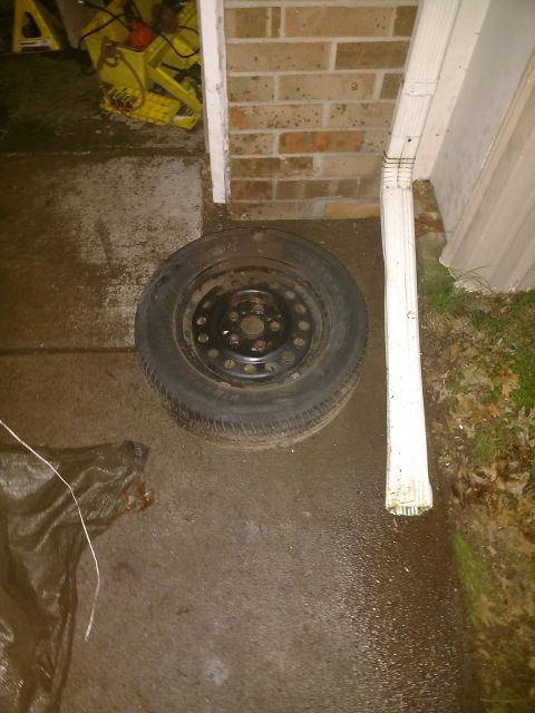 1989 Ford Probe - A spare tire is shown lying on a wet paved surface next to a downspout and a brick wall. A dark tarp is partially visible in the lower left…