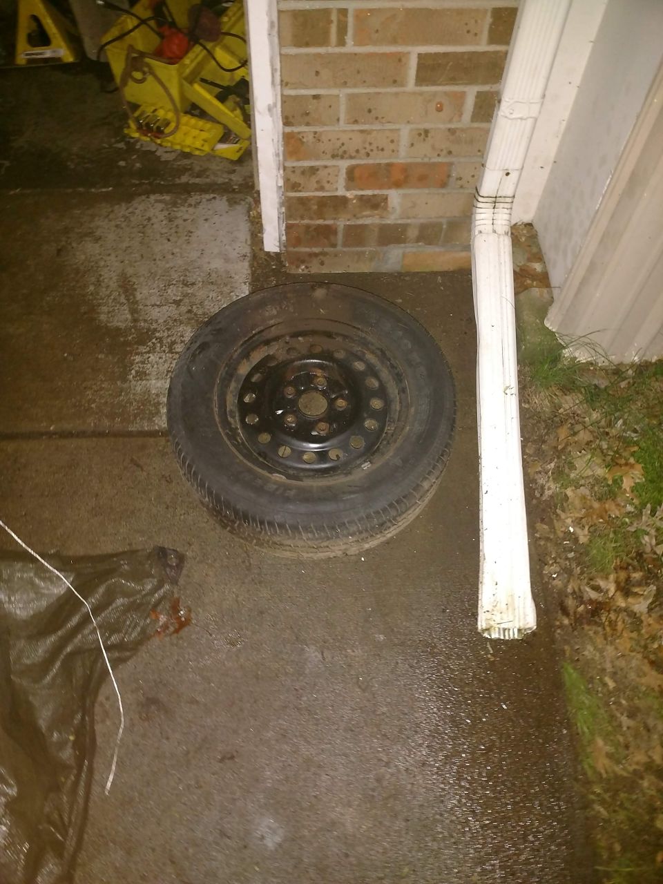 Spare tire on wet pavement next to a downspout and brick wall.