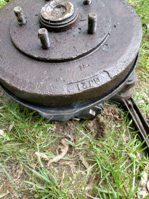 1989 Ford Probe - A close-up, low-angle shot of a rusty, dark-colored brake drum with visible lug studs, resting on grass and dirt. The drum has some engrave…