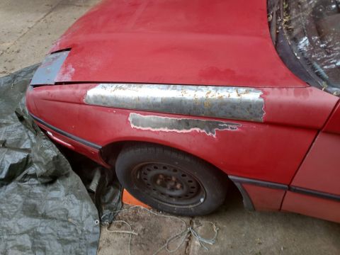 1989 Ford Probe - The front fender and hood of a red car are shown, with areas of rust and primer visible along a trim line. A dark green tarp is draped over…