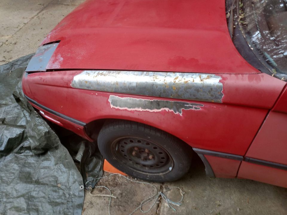 Red car fender and hood with rust and primer, partially covered by a green tarp.