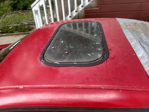 1989 Ford Probe - The red roof of a car is shown, featuring a sunroof with a dark trim and a slightly dirty glass panel. A portion of a tarp is visible on th…
