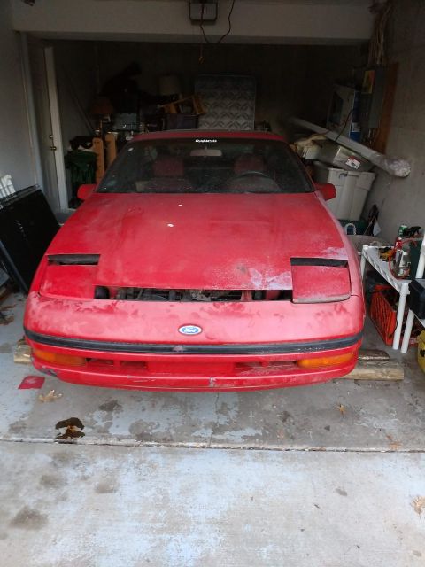 1989 Ford Probe - Front view of a red, dusty 1989 Ford Probe parked in a garage. The car is missing its grille and has a sticker on the windshield that reads…