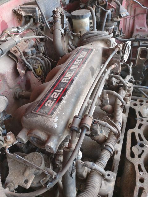 1989 Ford Probe - A close-up, overhead view of a dirty, gray engine intake manifold with red and black "2.2L FUEL INJECTION" text. Various hoses, wires, and…