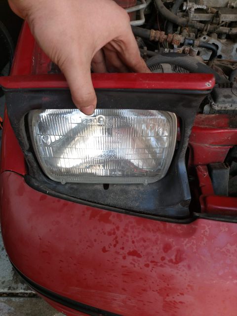 1989 Ford Probe - A hand is shown holding the edge of a red car's headlight assembly. The headlight lens is clear with visible internal elements and text, an…