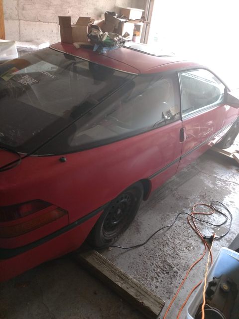 1989 Ford Probe - The rear passenger side of a red 1989 Ford Probe is shown in a garage. The car has a sunroof and a sticker on the rear window that says "BE…