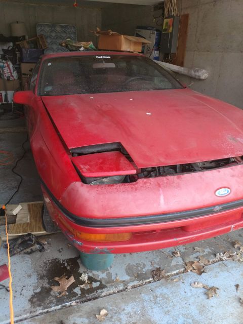 1989 Ford Probe - A red Ford Probe is parked in a garage, with its hood open and the front bumper showing some wear and tear. The car is covered in dust, sug…