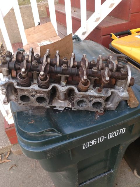 1989 Ford Probe - A disassembled engine cylinder head with visible rust sits on top of a dark green trash bin. The cylinder head has multiple valves and rock…