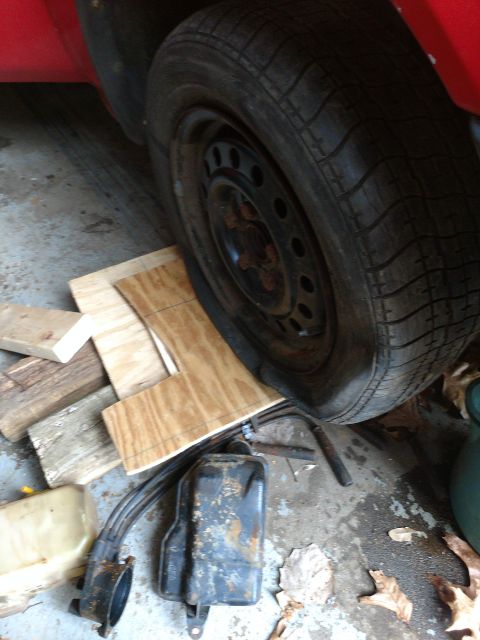 1989 Ford Probe - A close-up view shows the front passenger side tire and wheel of a red vehicle, resting on wooden planks on a concrete floor. Various car p…