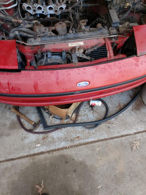 1989 Ford Probe - The front of a red Ford Probe is shown with the hood open, revealing the engine bay and radiator. Various tools and car parts are laid out…