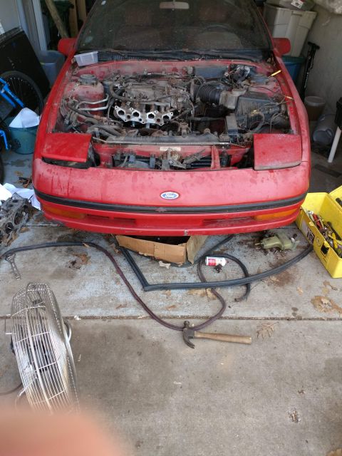1989 Ford Probe - A red Ford Probe with its hood open, revealing the engine bay. Tools and car parts are scattered on the concrete ground around the vehicle.