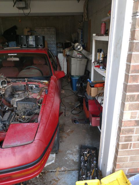 1989 Ford Probe - The front driver's side of a red 1989 Ford Probe is visible, with its hood open and engine compartment exposed. The car is parked in a clut…