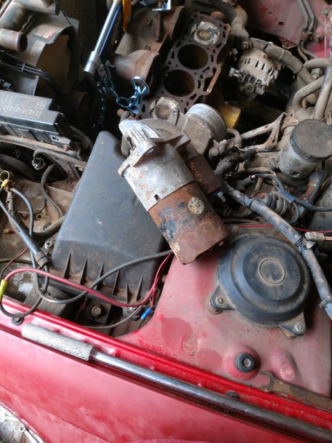 1989 Ford Probe - A close-up view of a rusty starter motor and other engine components in a red vehicle's engine bay. A chain is looped around some of the pa…