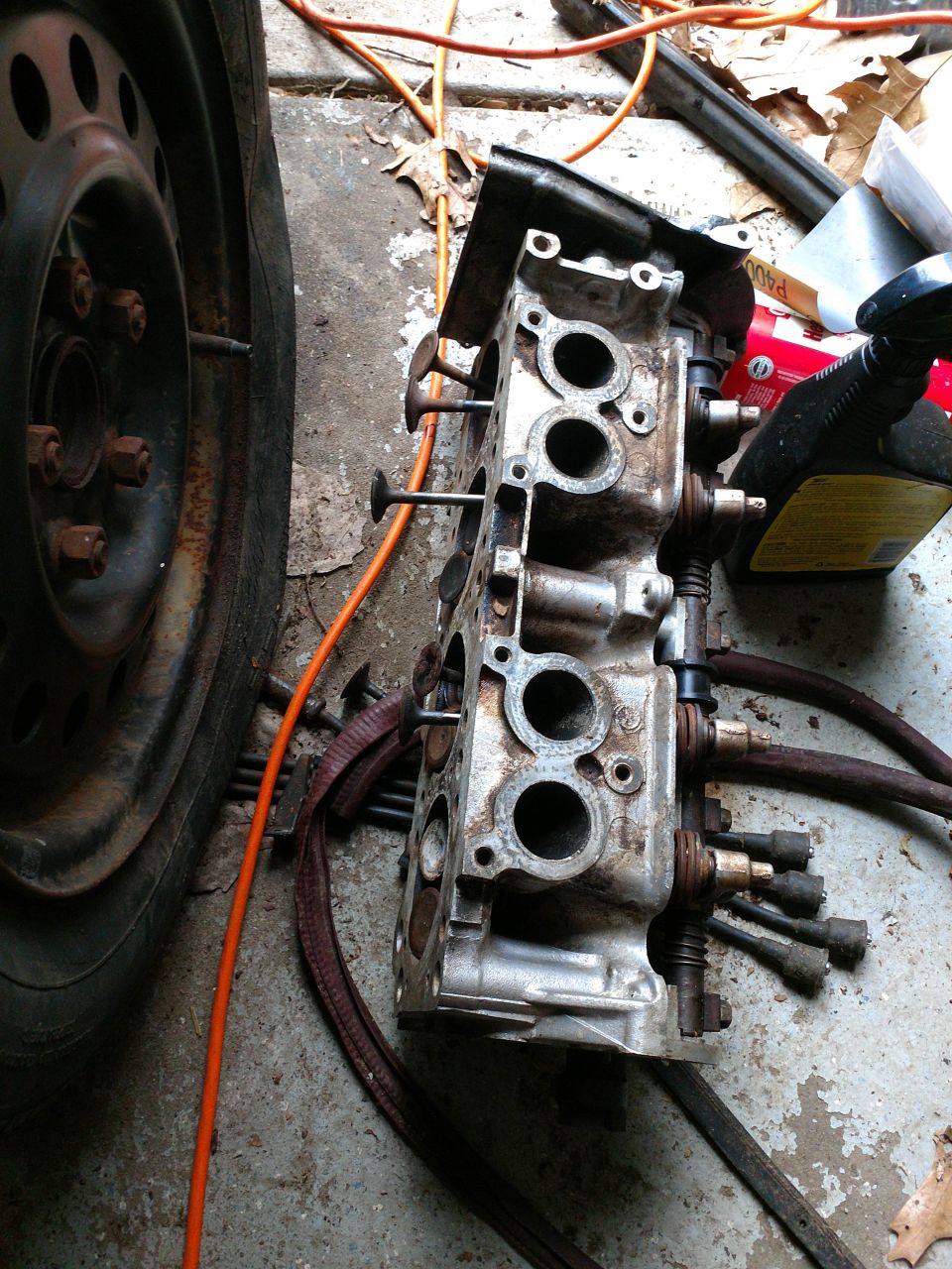 Disassembled engine cylinder head with spark plug wires next to a car tire.