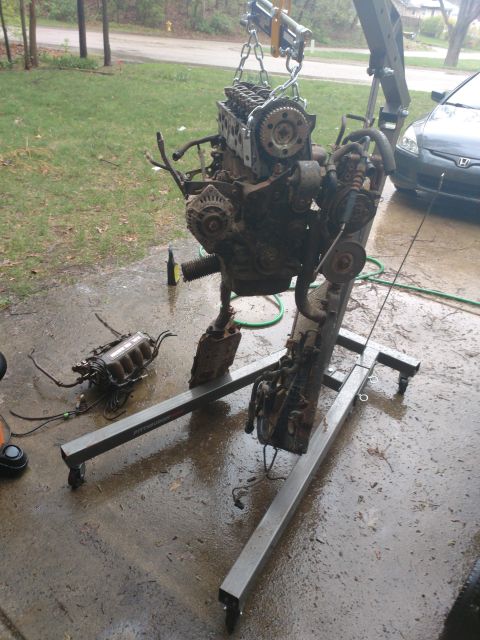 1989 Ford Probe - A disassembled engine is suspended by a chain hoist attached to an engine stand. Another engine component rests on the wet concrete ground…