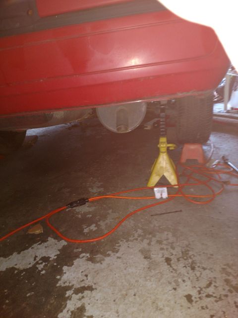 1989 Ford Probe - The rear underside of a red car is supported by a yellow jack stand. An orange extension cord runs across the concrete floor beneath the ve…