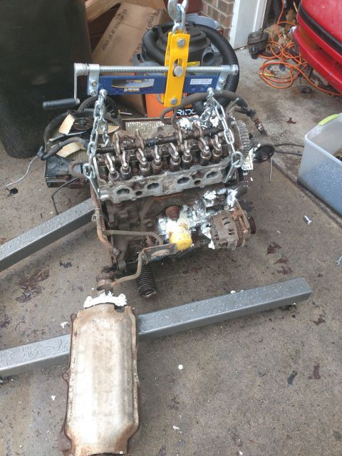 1989 Ford Probe - A disassembled engine is suspended by a yellow engine hoist with chains attached to the engine block. A catalytic converter is visible in t…