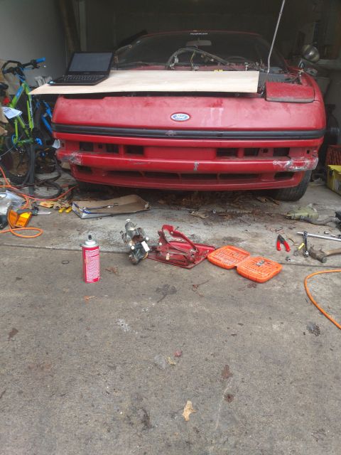 1989 Ford Probe - Front view of a red Ford car with its hood open and a piece of wood covering the engine bay. Various tools and car parts are scattered on t…