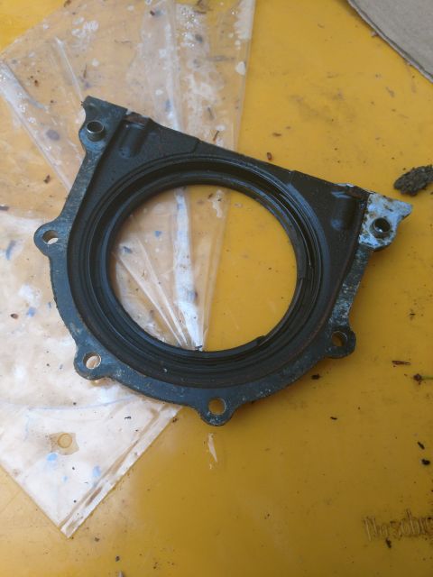 1989 Ford Probe - A black oil seal or gasket with a circular opening is shown on a yellow surface. The part has several bolt holes around its perimeter and a…