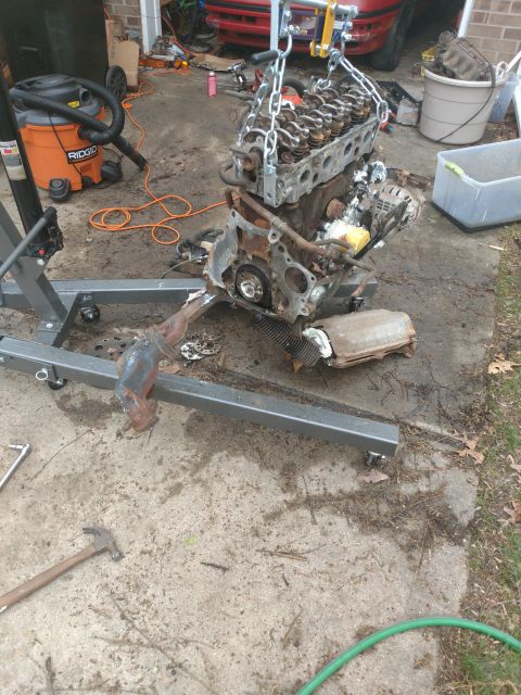 1989 Ford Probe - A disassembled engine is suspended by chains from an engine hoist, resting on a concrete driveway next to a shop vacuum. Various engine com…