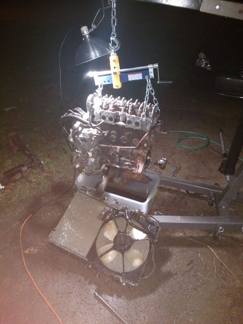 1989 Ford Probe - An engine is suspended by a chain hoist and engine support bar, with a radiator and fan assembly lying on the wet ground below. A work ligh…